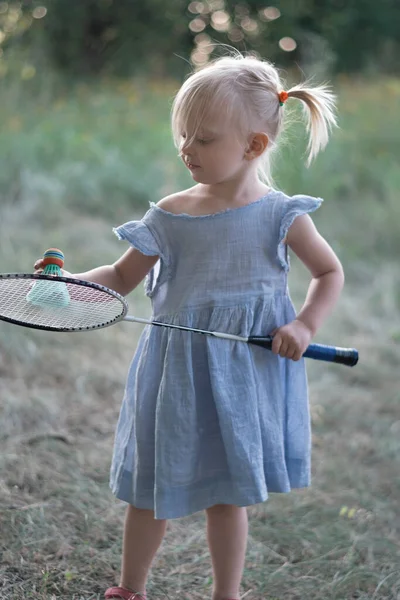 Three-year-old child plays badminton. Cute little girl in summer blue dress holds badminton racket. Vertical frame.