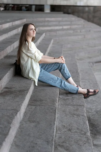 Portrait of girl sitting on the stairs. Modern young woman in jeans and shirt resting outside on the stairs