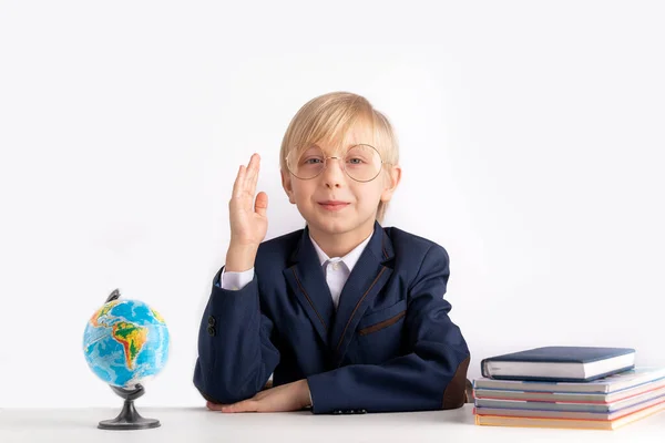 Schoolboy in large glasses sits at table with textbooks with his hand raised. Excellent student. Boy knows the answer