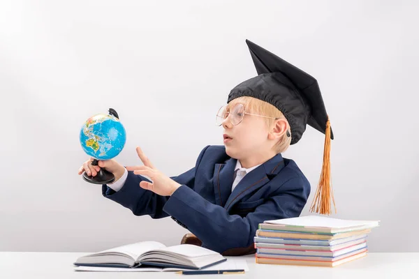 Boy in school uniform and students hat with globe in his hands near books and notebooks.Schoolboy studies geography.