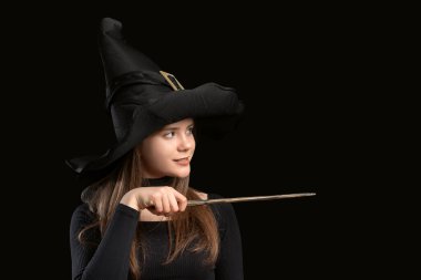 Young girl in witch costume wears pointed hat, black clothes and uses magic wand. Portrait isolated on black background. Copy space