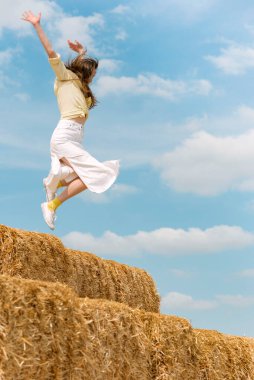 Woman while jumping on big haystack. Girl has fun and jumps in the hay on blue background. Summer vacation.