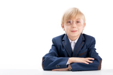 Elementary school student wears school uniform and glasses sit idly by at desk. Portrait of blond schoolboy on white background. Copy space