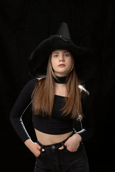 Girl in black clothes and pointed hat on black background. Vertical frame. Modern witch. Halloween