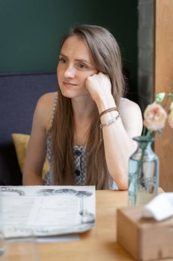Pensive young woman sits in a cafe with her head resting on her hand. Tired look to nowhere.