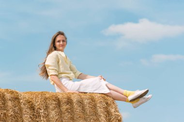 Young pretty woman sitting on haystacks on blue sky background. Girl resting on bale of hay in summer field