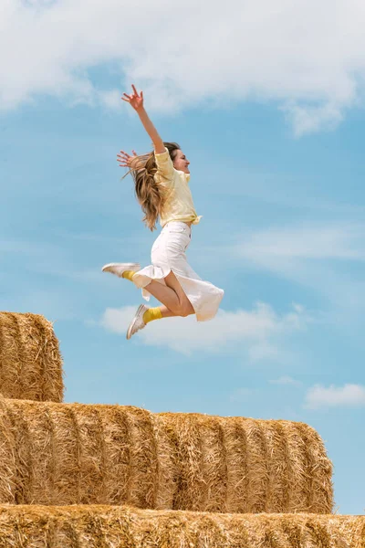 Young woman jumps on the haystack with her hands raised high. Countryside holiday on the farm, blue sky. Hay harvesting