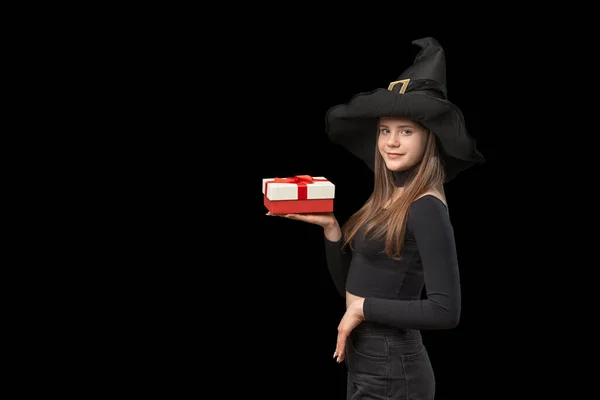 Cute young woman in witch black hat with gift on palm. Girl on black background. Copy space.