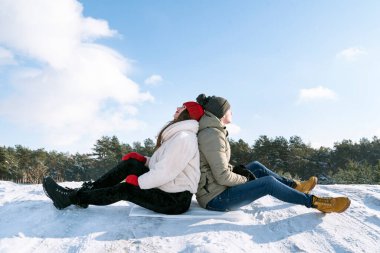 Guy and girl sit back to back in the snow. Active winter recreation.