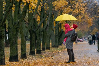 Anne, bir sonbahar parkında sarı yapraklardan oluşan bir arka planda sarı bir şemsiye altında duran küçük bir çocuğu kollarında tutuyor..