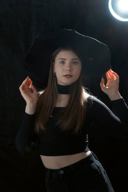 Siyah elbiseli ve geniş kenarlı şapkalı genç bir kadının portresi. Gotik tarzda genç bir kız. Stüdyo ışığı. Dikey çerçeve. Siyah arkaplan