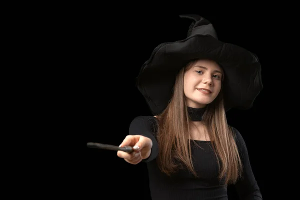 魔女の肖像画は黒い服と尖った帽子を身に着けている 少女は魔法の杖を使う 黒の背景に隔離 ハロウィン衣装 — ストック写真