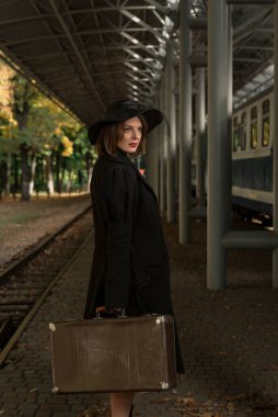 Eski moda valizli genç bir bayan tren istasyonunda bekliyor. Paltolu ve şapkalı orta yaşlı kadın portresi