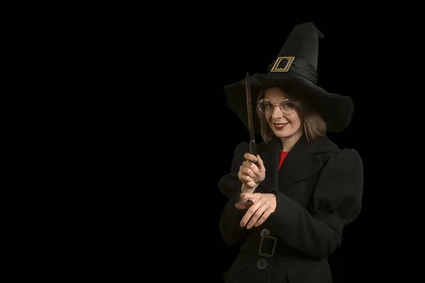 Young woman in form whitch in black hat and with magic wand isolated on black background. Costume party.