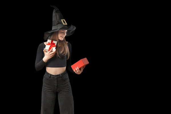 Smiling girl in black clothes and pointed hat holds an open gift. Looks at box in surprise. Witch girl isolated on black background