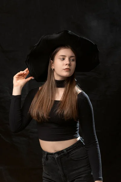 Beautiful young lady wears black clothes and wide-brimmed hat. Portrait of young model on black background. Vertical frame