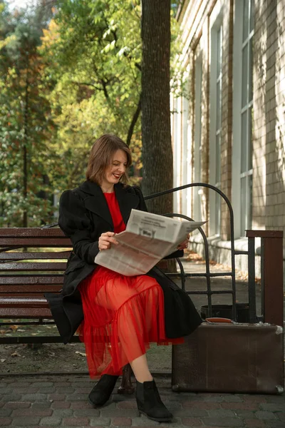 Joven Alegre Vestido Rojo Abrigo Negro Sienta Banco Leyendo Periódico — Foto de Stock