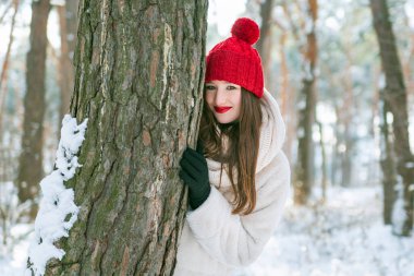 Kırmızı şapkalı ve eldivenli güzel esmer kadın karlı bir parktaki bir ağaca sarılıp yatıyor. Açık havada portre.