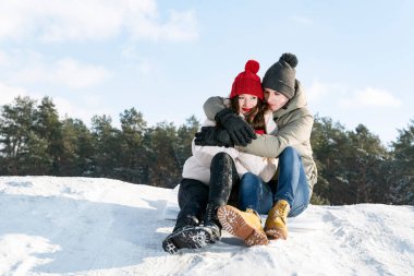 Genç bir adam ve bir kadın, ormanın ve mavi gökyüzünün arka planında karda oturup sarılır. Birkaç tane de açık havada. Boşluğu kopyala.