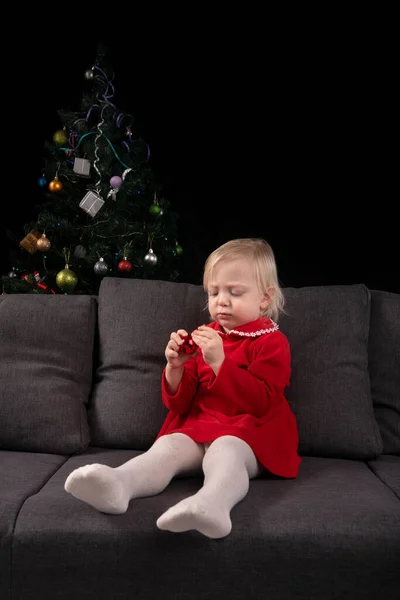 little girl sits on a sofa against the background of a Christmas tree and looks at a Christmas tree toy. New Year eve. Vertical frame.
