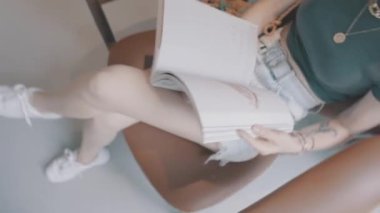 Blond hair girl is sitting on the brown armchair and reading magazine 4K footage
