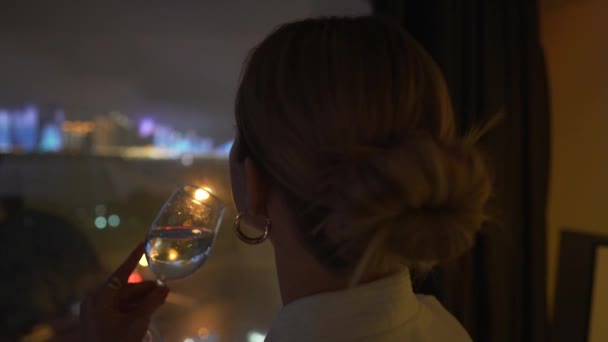 穿着白色浴衣的金发小女孩站在窗边 喝着酒杯里的香槟 看着夜市灯火通明的4K镜头 邻近城市景观 夜间大都市 亚洲城市 — 图库视频影像