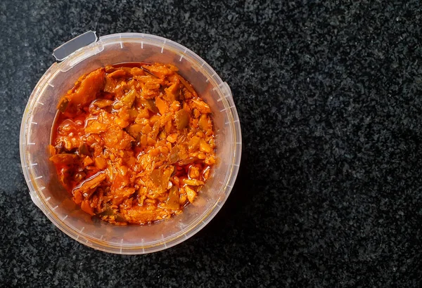 close up top view on a plastic bucket container filled with spicy mango achar which is a type of food made from unripe green mangoes pickled in oil with different types of spices.