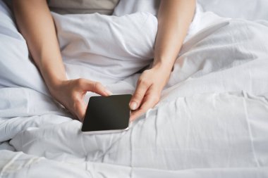 Beyaz yatakta telefonu kullanan bir kadının eli. Kesme yolu.