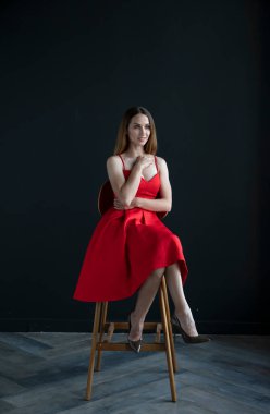 Koyu arkaplanda kırmızı elbiseyle oturan güzel genç bir kadının portresi.