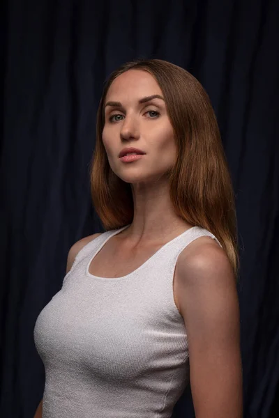 Portrait of determined young woman wearing white top looking at camera posing on dark background