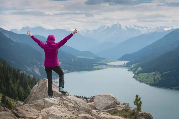 a sporty woman looking into the distance and rising her arms seen from behind while hiking on a prominent vantage point over a wide valley with an elongated large lake in south tyrol