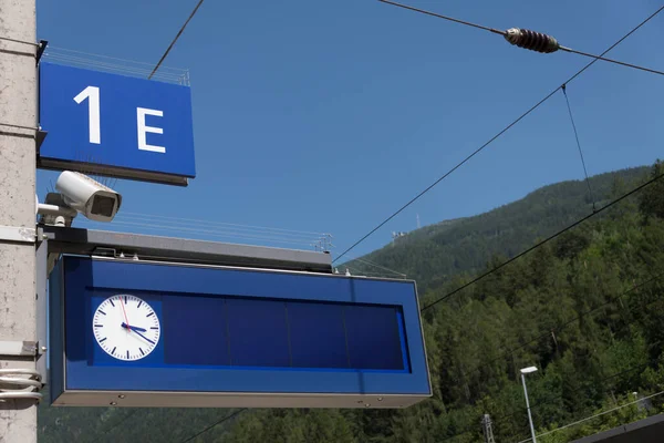 铁路车站带有模拟时钟的蓝色显示屏图像 带有摄像头监控和信息板 在蓝天的绿色植被前面有平台标识 — 图库照片
