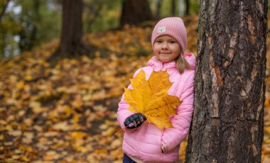 Bir kız elinde büyük sarı bir akçaağaç yaprağı tutuyor..