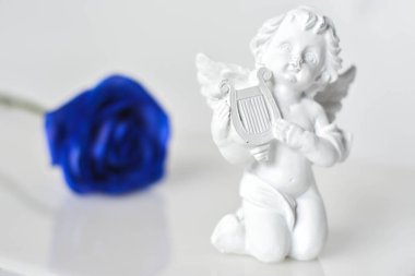Beyaz parlak yüzeyde seramik bir melek figürü ve arkasında mavi bir gül.