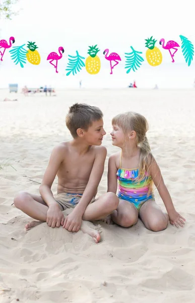 Маленька дівчинка і хлопчик сидять на пляжі з гірляндою, що зображує фламінго і ананаси Ліцензійні Стокові Фото