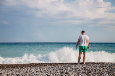 attractive young adult man stands with his back on the shore of the mediterranean sea with waves. copy space.