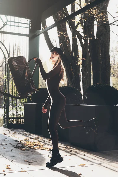 Modische junge brünette Frau mit langen Beinen trägt solide langärmelige Bodycon One Piece Jumpsuits und posiert mit einer ledernen braunen Reisetasche — Stockfoto