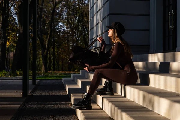 Modische junge brünette Frau mit langen Beinen trägt solide langärmelige Bodycon One Piece Jumpsuits und posiert mit einer ledernen braunen Reisetasche — Stockfoto