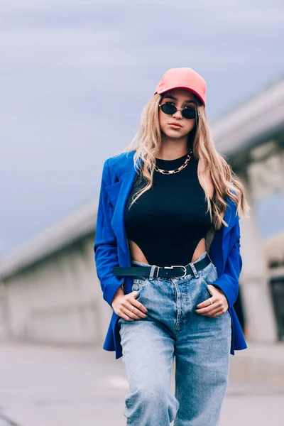 Sokakta poz veren genç seksi sarışın hippi kadın. Mavi bir ceket, kot pantolon, beyzbol şapkası ve güneş gözlüğü takıyor.. — Stok fotoğraf
