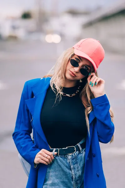 Junge sexy blonde Hipsterfrau posiert auf der Straße. Mit blauer Jacke, Jeans und Baseballmütze und Sonnenbrille. — Stockfoto