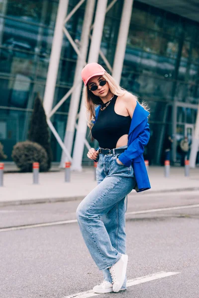 Jovem mulher hipster loira sexy posando na rua. Vestindo casaco azul elegante, jeans e chapéu de beisebol e óculos de sol. — Fotografia de Stock