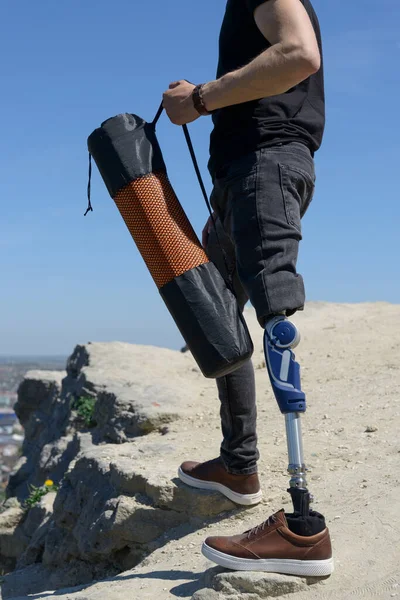 Um homem numa perna protética viaja pelas montanhas. Vestido com jeans pretos e uma camiseta, ele carregando tapete — Fotografia de Stock