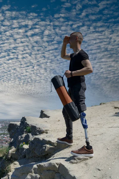 Um homem numa perna protética viaja pelas montanhas. Vestido com jeans pretos e uma camiseta, ele carregando tapete — Fotografia de Stock