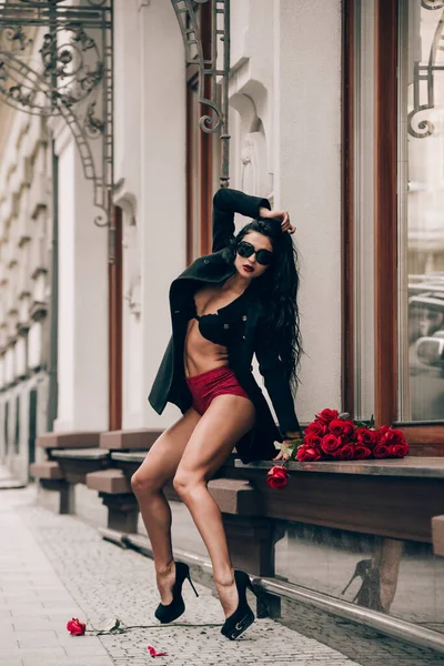 Κομψό όμορφη μελαχρινή γυναίκα με γυαλιά ηλίου και κόκκινο σορτς και μαύρο παλτό κορίτσι που θέτουν κάθεται στο περβάζι του παραθύρου με ένα μεγάλο μπουκέτο κόκκινα τριαντάφυλλα. Γενέθλια. Ημέρα του Αγίου Βαλεντίνου. Επιλογή εστίασης, filmgrain — Φωτογραφία Αρχείου