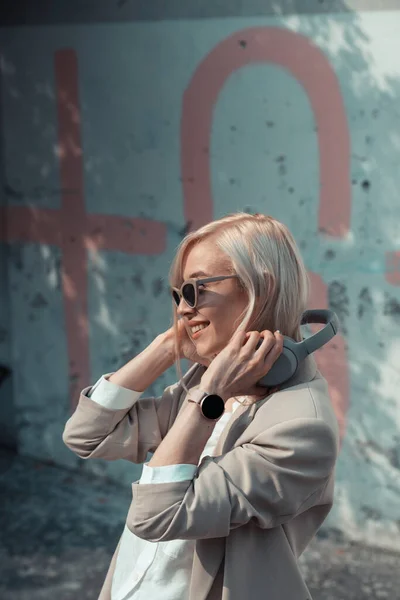 Portrait of blonde woman with headphones in hands. Hipster fashion model wear stylish wireless headphones enjoy listen new cool music. — Stock Photo, Image