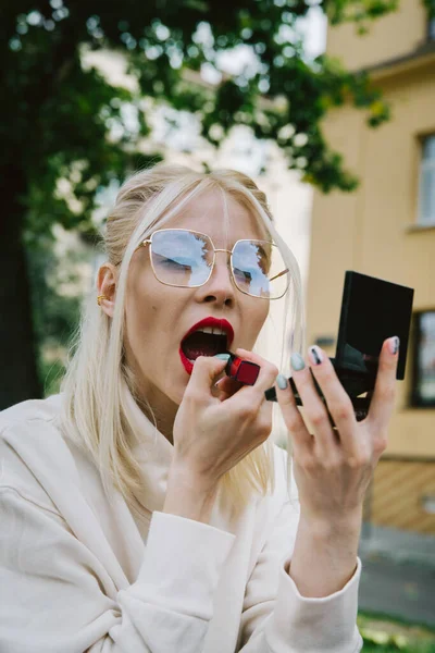 Young smiling hipster happy woman doing make-up using lipstick and mirror outdoors — Stock Photo, Image