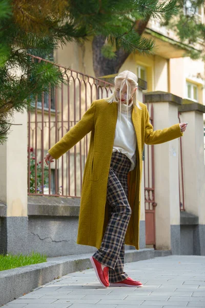 Mujer joven hermosa de moda con el pelo rubio en un abrigo largo con estilo, pantalones a cuadros, zapatos rojos y gafas posa en las calles de la ciudad. — Foto de Stock