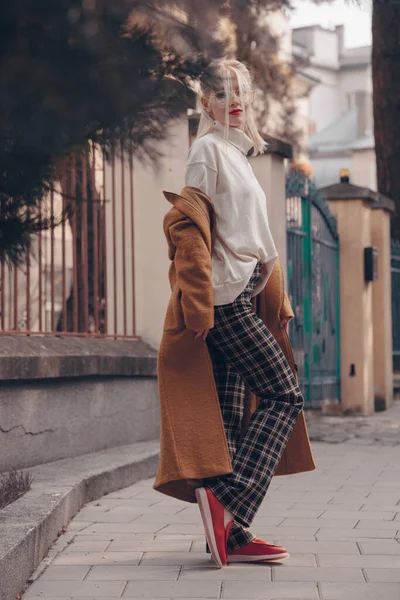 Mujer joven hermosa de moda con el pelo rubio en un abrigo largo con estilo, pantalones a cuadros, zapatos rojos y gafas posa en las calles de la ciudad. — Foto de Stock