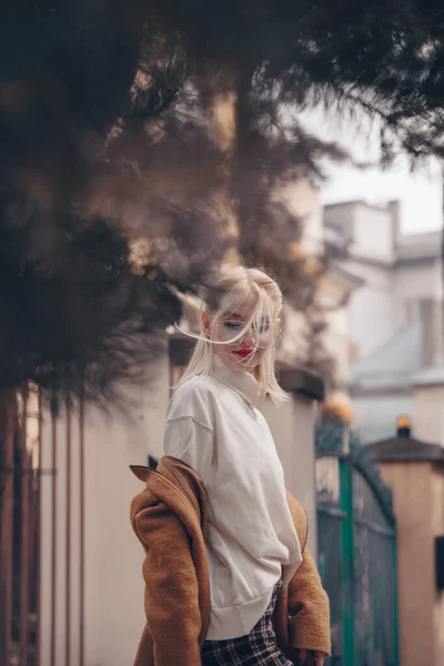 Bella donna alla moda con i capelli biondi in un cappotto lungo elegante, pantaloni a scacchi e occhiali posa nelle strade della città. — Foto Stock