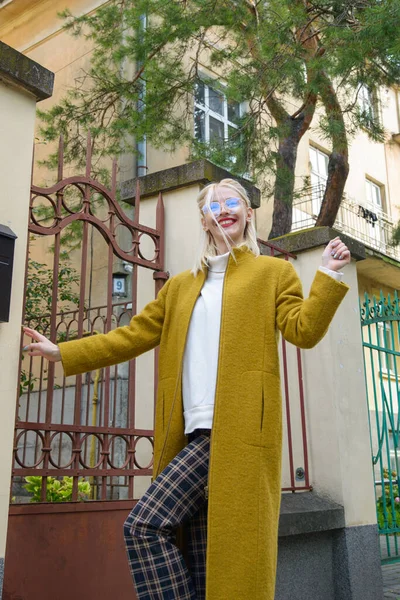 Mujer joven hermosa de moda con el pelo rubio en un abrigo largo con estilo, pantalones a cuadros y gafas posa en las calles de la ciudad. — Foto de Stock